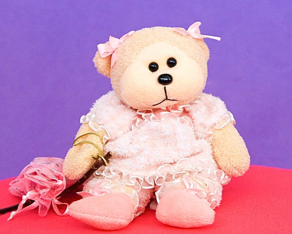 Frilly, a small beanie baby teddy in a frilly romper suit sits down on a pink cushion